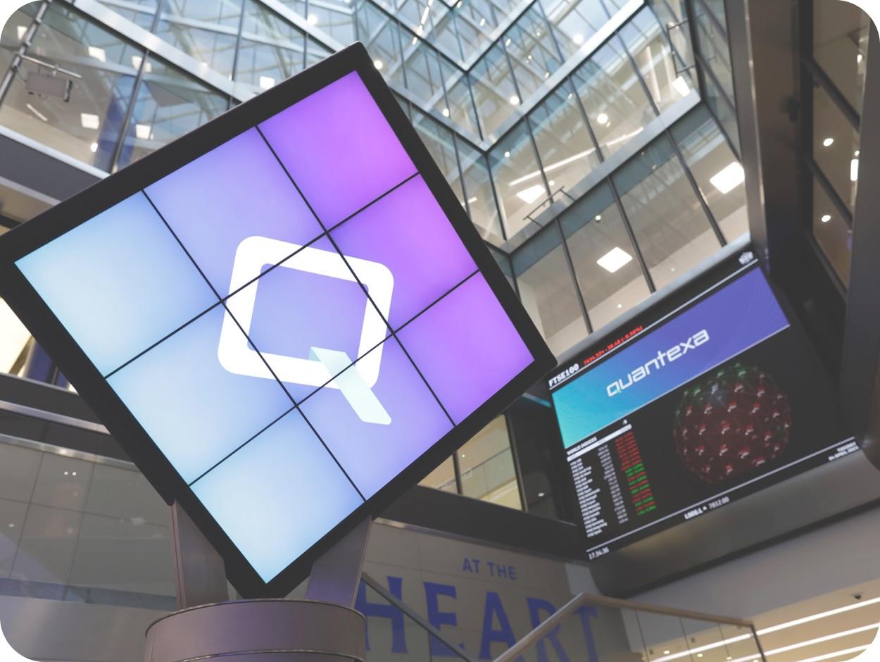 Digital display with a logo on a multi-screen setup in a modern building interior, alongside a stock market ticker showing "quantexa."