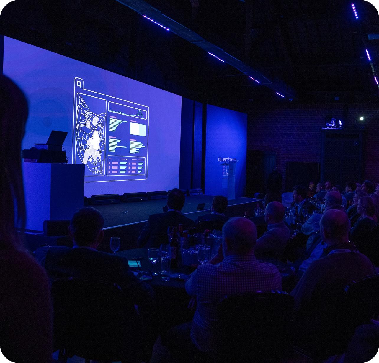 Audience watching a presentation with blue lighting in a dark room, featuring a large screen displaying digital graphics and text.