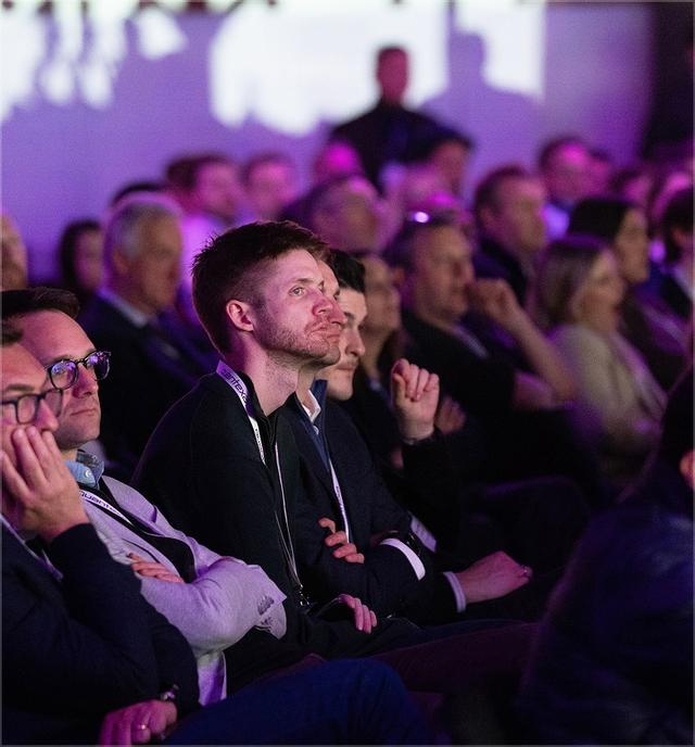 Audience members attentively watching a presentation, seated closely in a dimly lit venue with purple ambient lighting.