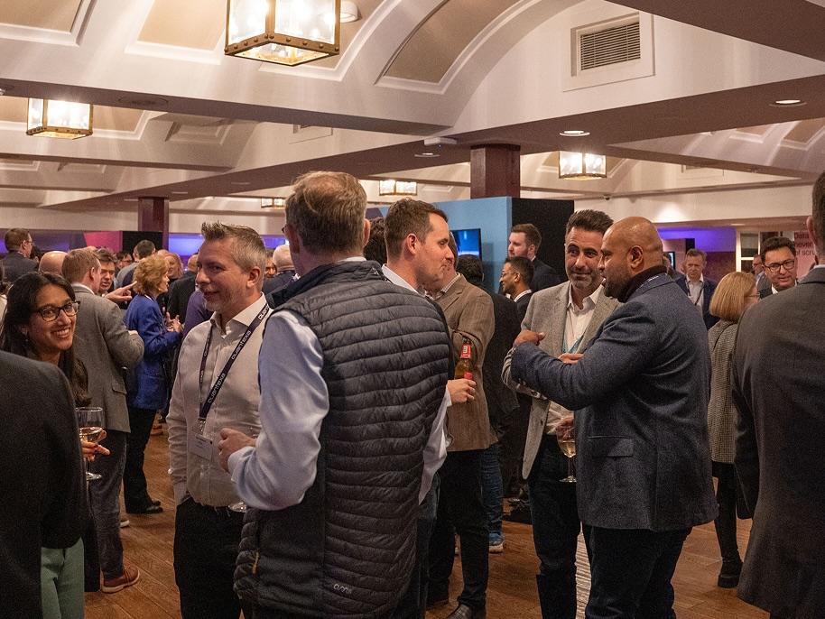 People mingling at a networking event in a large room with arched ceilings and chandeliers. Some hold drinks, engaging in conversations.