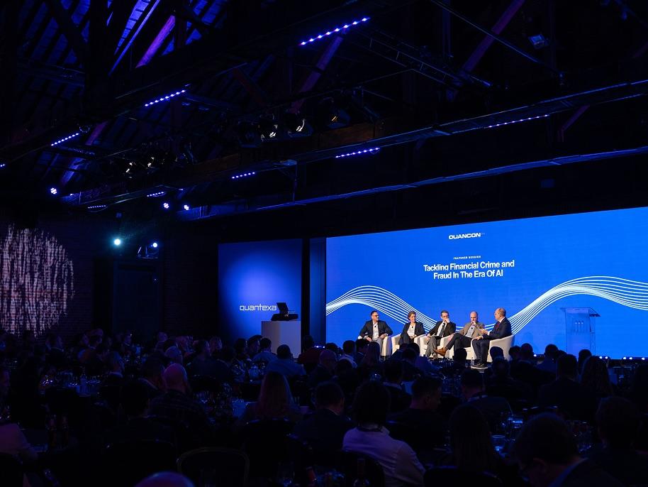 Panel discussion on stage at a conference, with four speakers seated under blue lighting, and an audience in a dimly lit room.