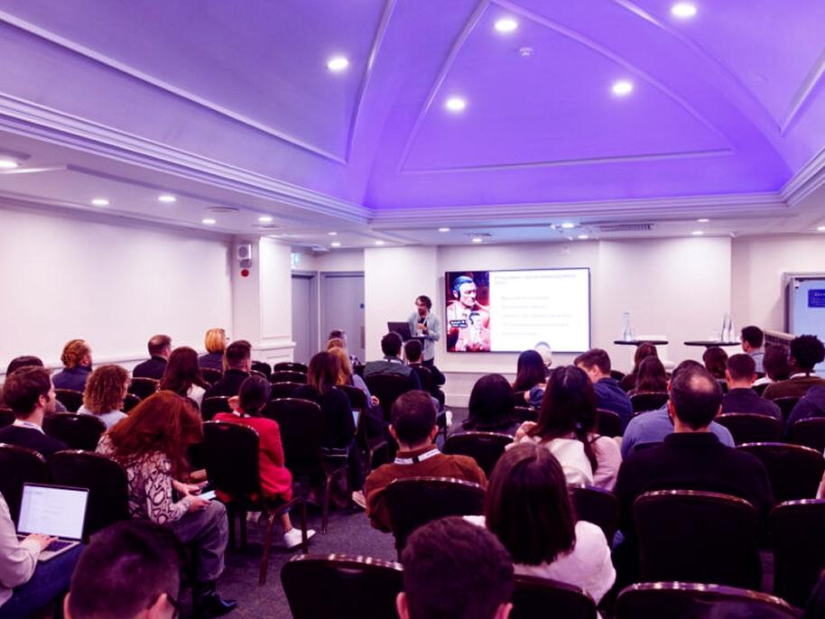 A speaker addresses an audience in a conference room with a purple-lit ceiling. A presentation slide is displayed on a large screen.