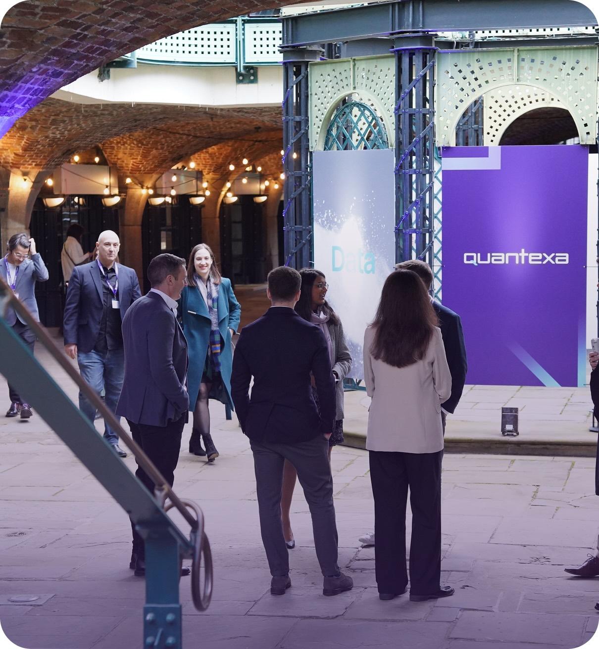 People in business attire gather at an indoor event with "Quantexa" and "Data" banners, under a brick arch ceiling with lights.