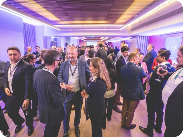 A large group of people in formal attire engage in conversations at a networking event in a well-lit room with purple ambient lighting.