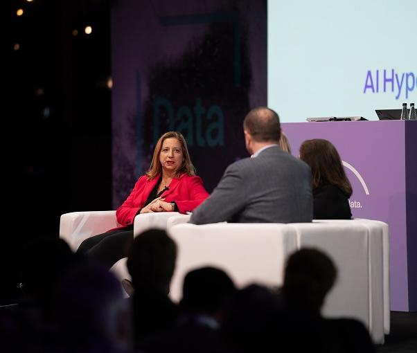A woman in a red blazer speaks on a panel with three other people, seated on white chairs at a conference.