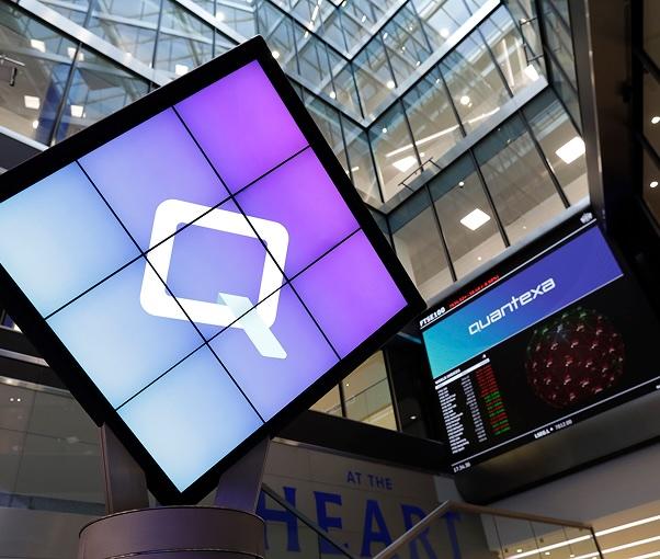 Digital display with a large "Q" logo in a modern building, next to a stock market screen showing "quantexa" and market data.