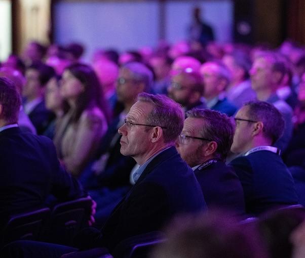 Audience members seated at a conference, attentively listening under purple lighting.