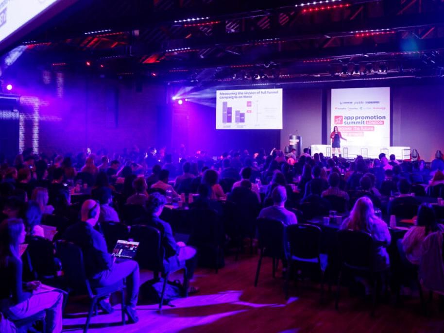 A large audience attends a conference in a dimly lit auditorium, with a speaker on stage and presentation slides projected on screens.