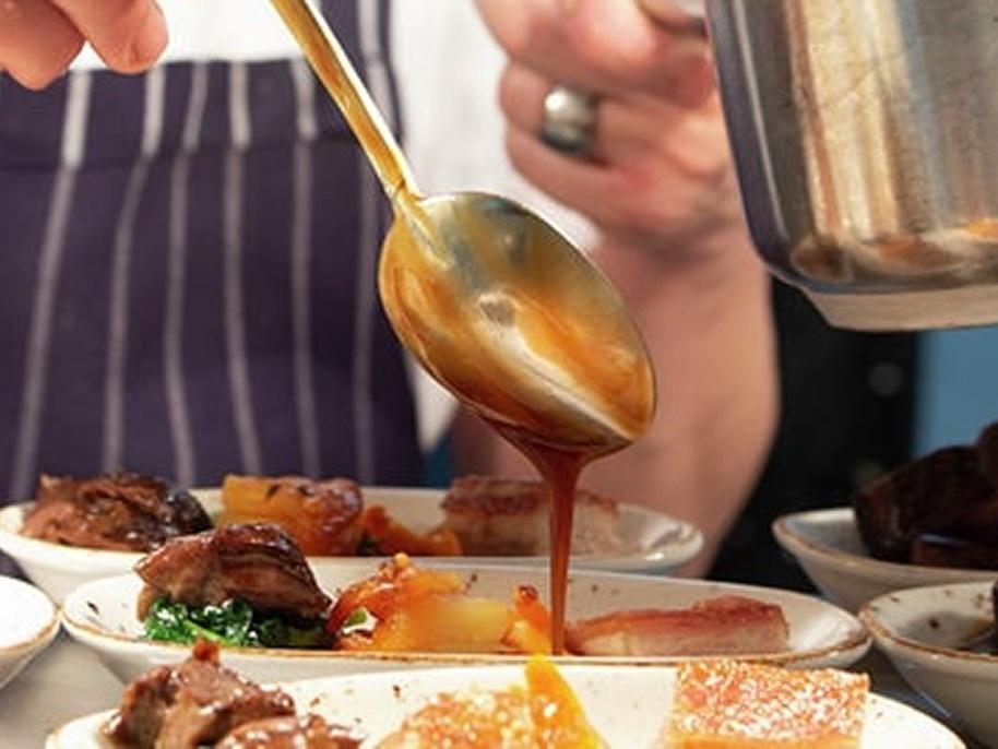Chef drizzling sauce over plated gourmet dishes with a ladle, wearing a striped apron, showcasing intricate food presentation.